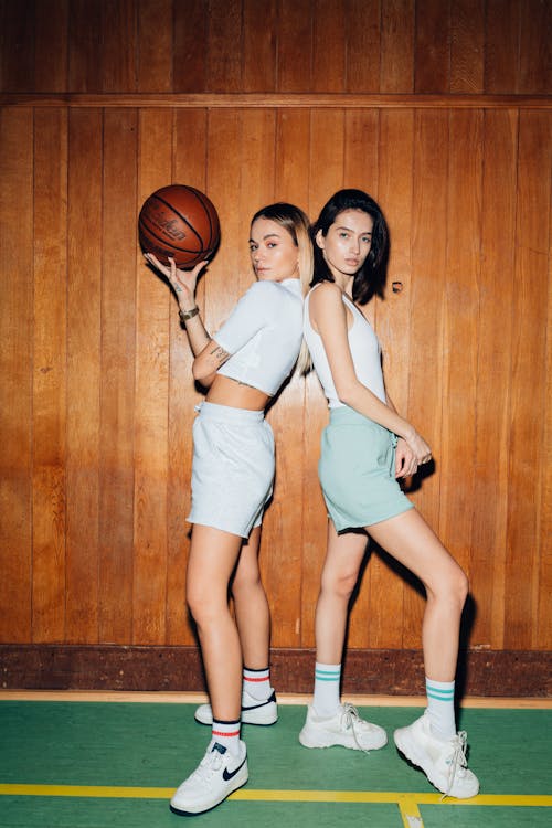 Two Young Women in White Tops and Shorts Posing at an Indoor Basketball Court