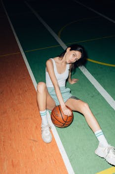 A young woman in a tank top sitting on a vibrant basketball court holding a basketball.