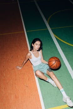 A young woman in a tank top sits on an indoor basketball court holding a basketball.