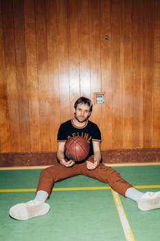 Man sitting on a gym floor holding a basketball in a relaxed, retro setting.