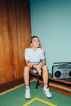 Young woman with tattoos sits indoors by a vintage boombox, embodying retro style.