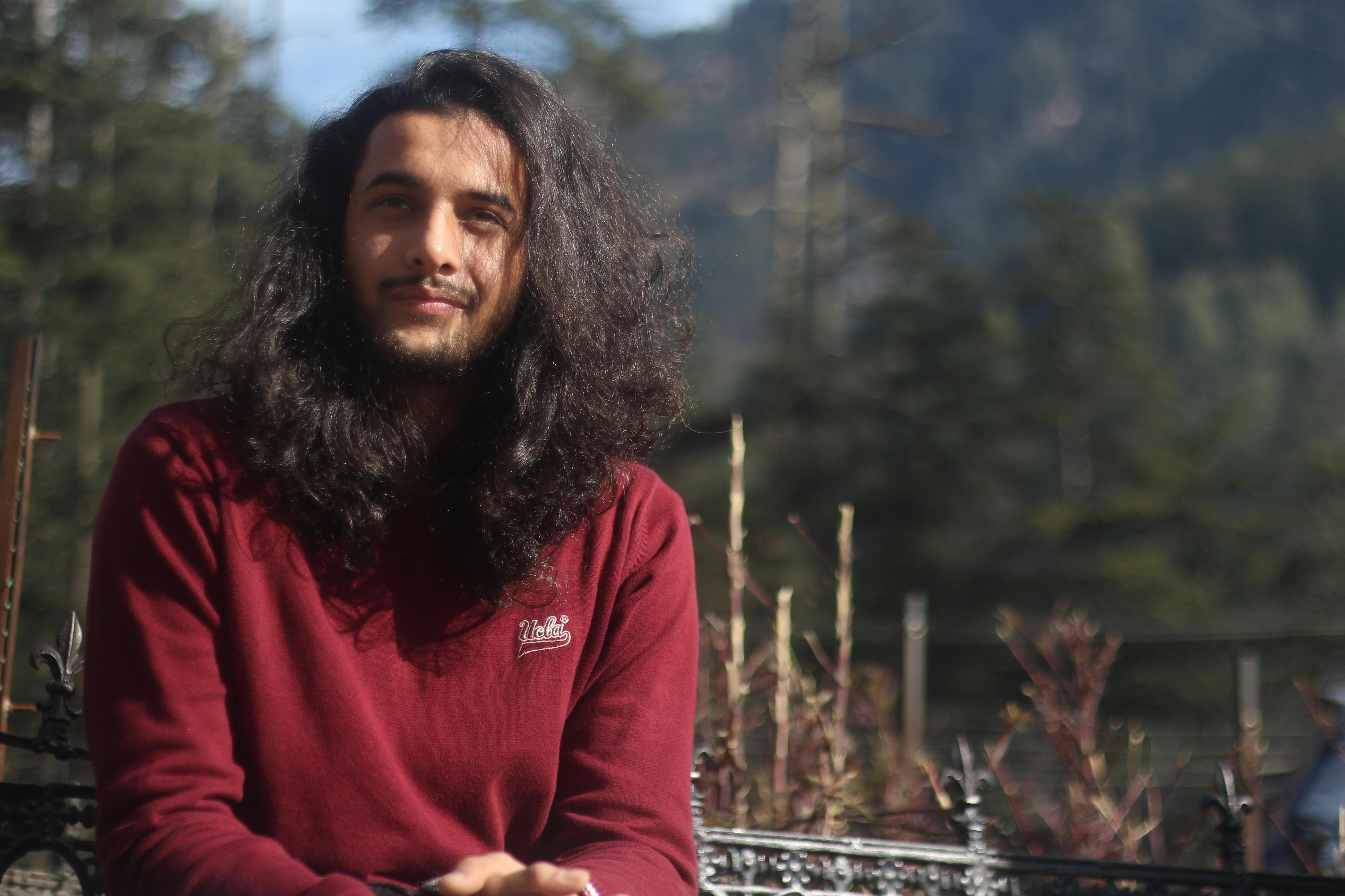 A relaxed outdoor portrait of a man with long hair and a beard wearing a red sweater.