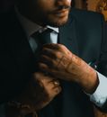 Crop anonymous well dressed ethnic male with beard and mehndi putting on tie during festive event