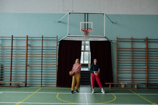 Two women holding basketballs in a gymnasium, standing by the hoop.