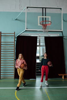 Two women in trendy sportswear holding basketballs indoors.
