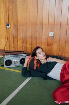 A woman in retro style clothing rests on a basketball beside a vintage boombox in an indoor setting.