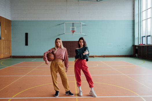 Two young women posing stylishly on a basketball court indoors.