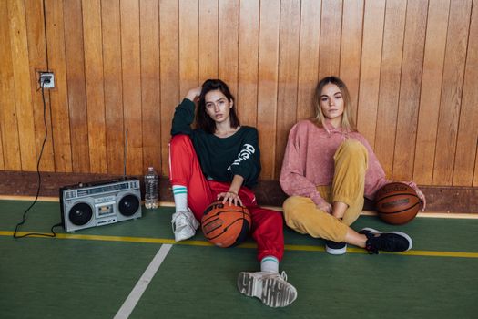 Two women in retro attire sit with basketballs and a boombox indoors.