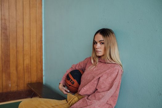 A young woman with a basketball sitting against a colorful wall indoors.