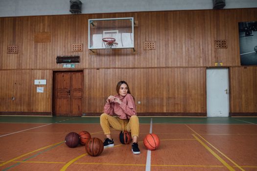 A young woman poses confidently in an indoor basketball court, surrounded by basketballs.
