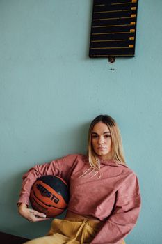 A young woman in a hoodie holds a basketball indoors against a teal wall.