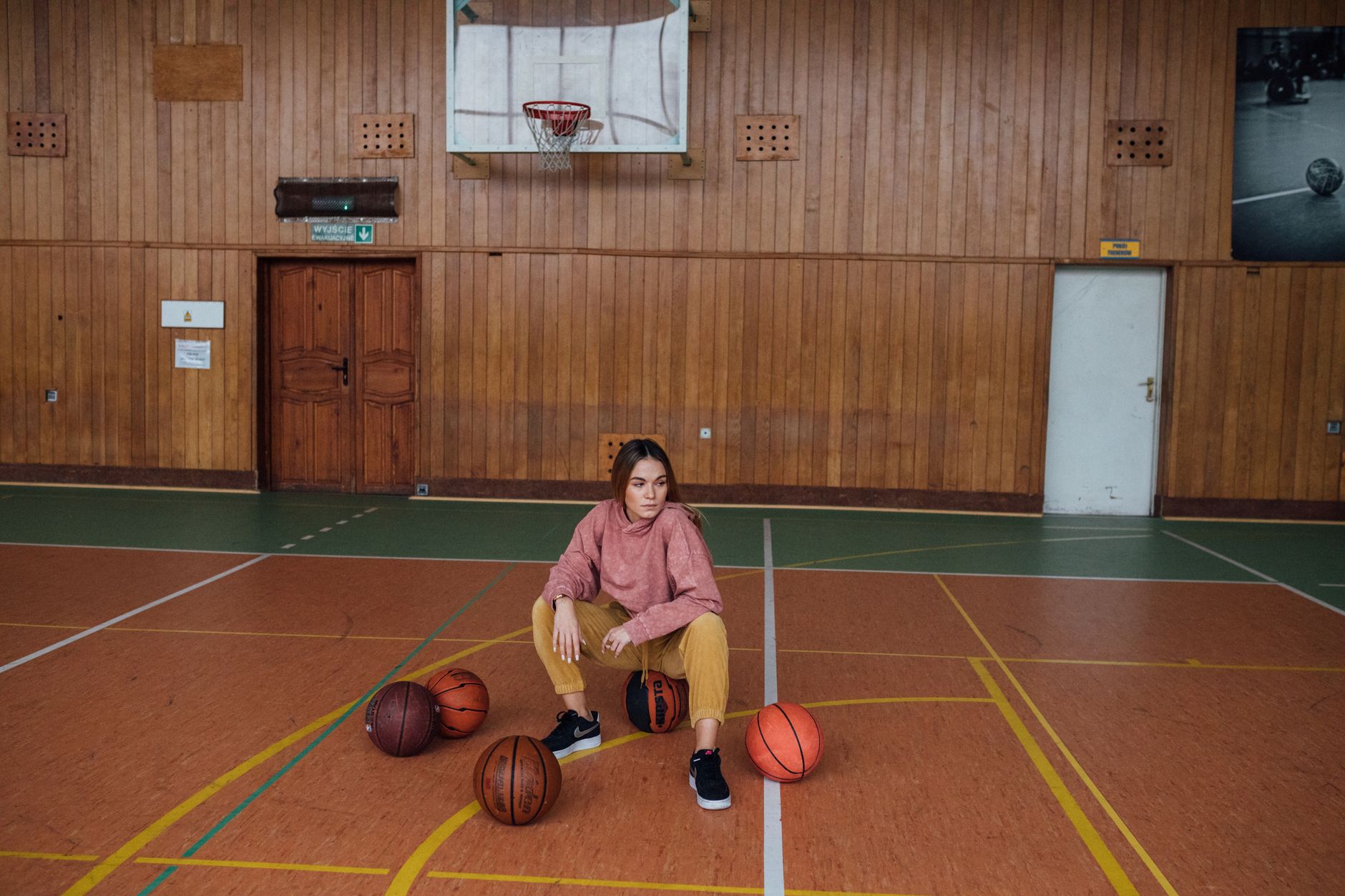 A woman in sporty attire sits on an indoor basketball court surrounded by basketballs.