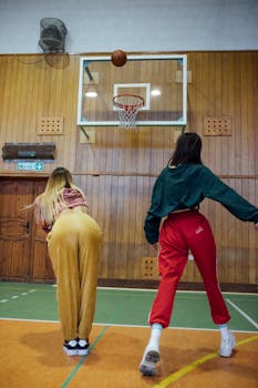 Two women playing basketball in an indoor gym with wooden walls.