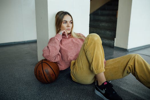Casual young woman in a hoodie sitting indoors, resting against a wall with a basketball.