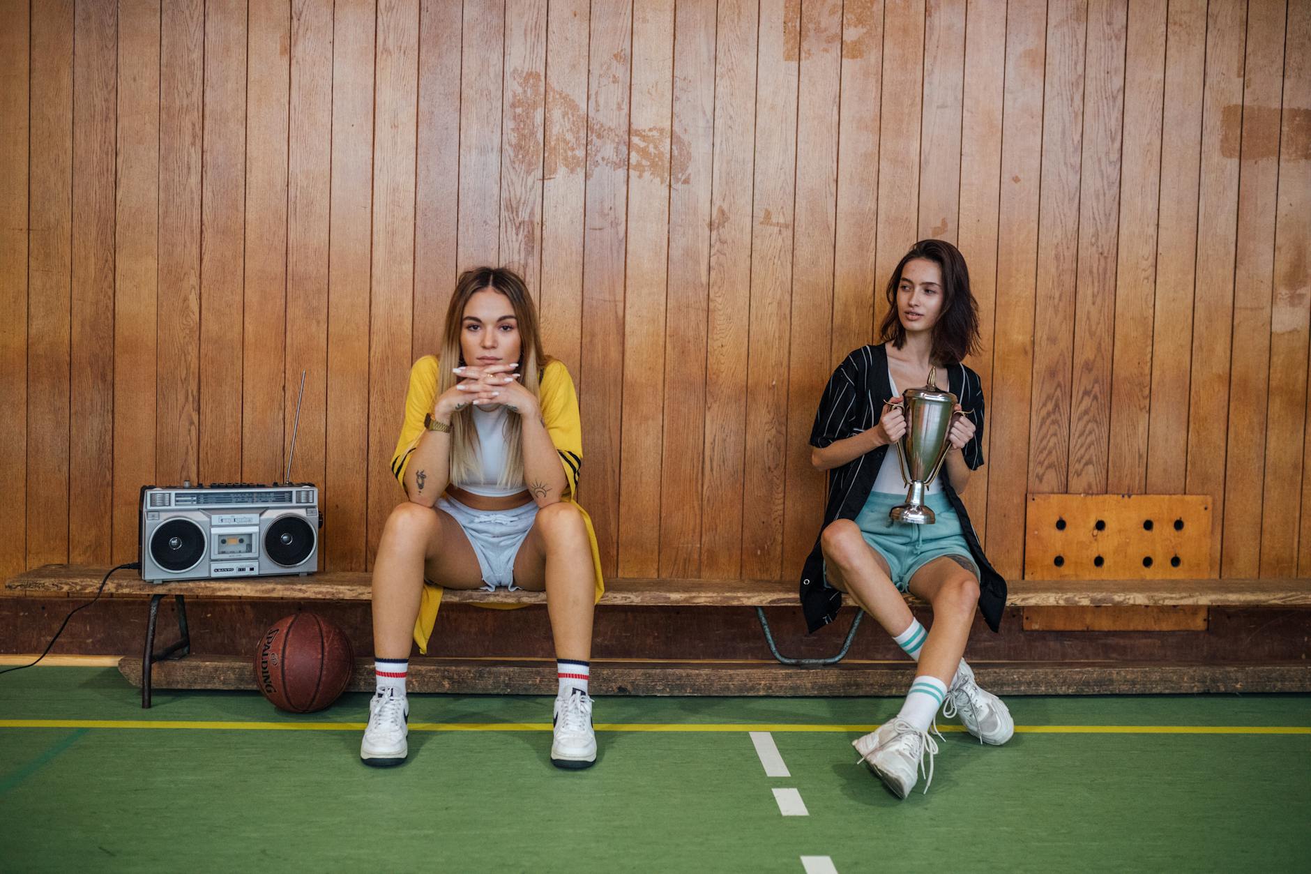 Two Women in Shorts and Long Shirts Sitting on a Bench at a Basketball Court