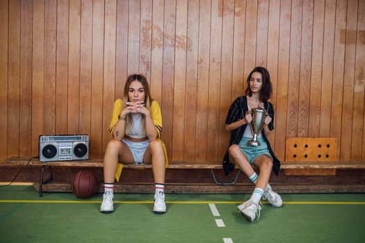 Two young women sitting on a bench with a trophy and a boombox, showcasing fashion in a sports setting.