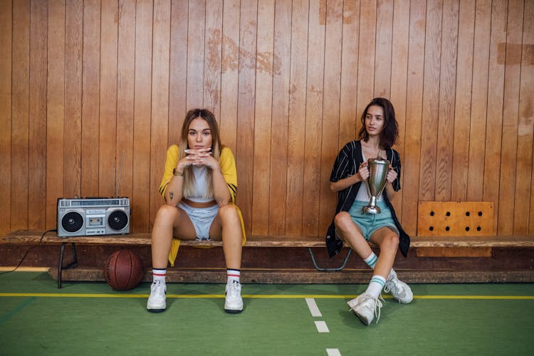 Two Women In Shorts And Long Shirts Sitting On A Bench At A Basketball Court