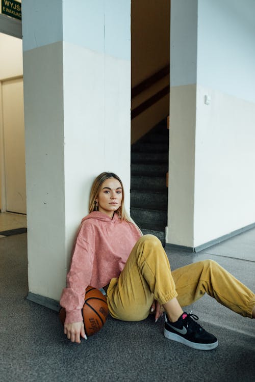 Blonde Young Woman in Pink Hoodie Sitting on Floor with Ball
