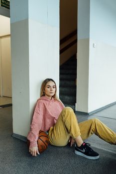 Stylish young woman in casual attire sitting indoors with a basketball and contemplative expression.