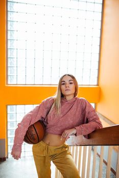 Trendy woman in casual fashion holding a basketball indoors, leaning on a stair railing.