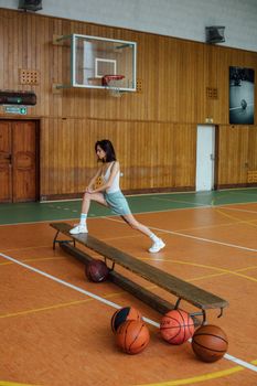 Caucasian woman stretching on a bench in an indoor basketball court with basketballs.
