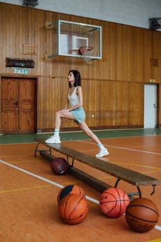 Woman in sportswear stretching on indoor basketball court with scattered basketballs.