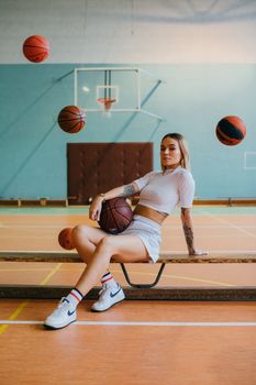Stylish woman sitting on a bench in a sports hall with basketballs around.