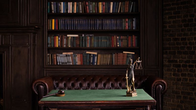 A classic study room with shelves of books, a gavel, and Lady Justice figurine on a green table.