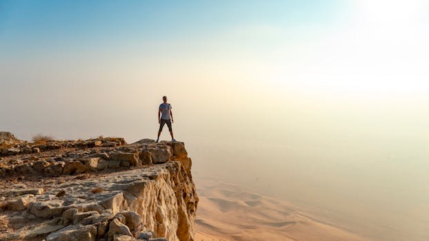 A man stands confidently on the edge of a rocky cliff overlooking a vast desert under a clear sky.