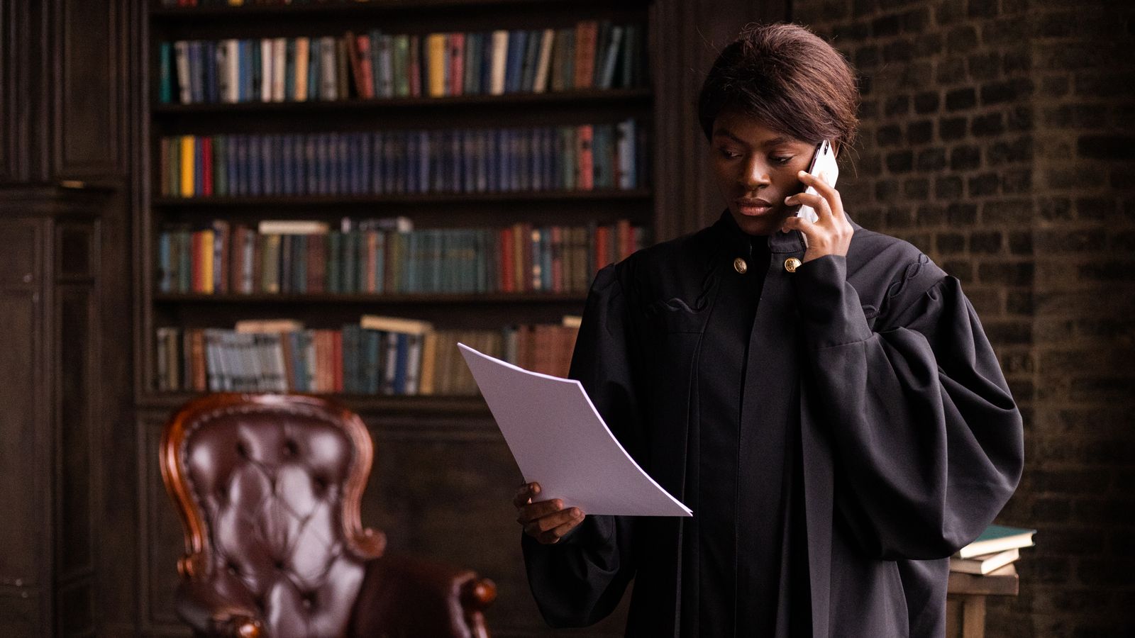 African American judge in traditional robes holds a document and talks on the phone in a law library.