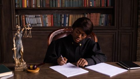 A judge in robes writing on a document at a desk in an office library with law books.