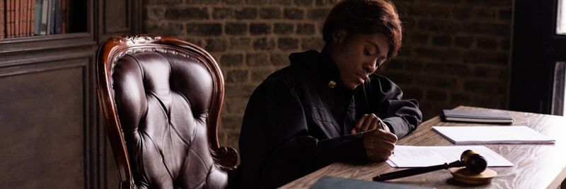 Black female judge in judicial robe writing at desk in an office setting, focusing on legal work.