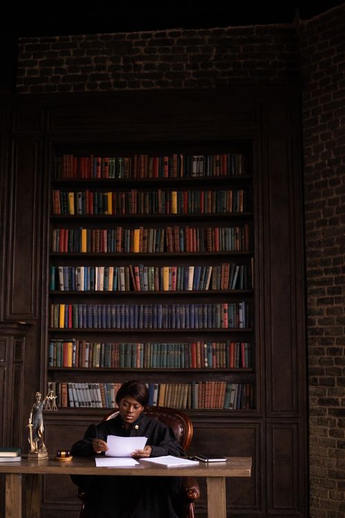 Free Black female judge reading papers at desk in office with bookshelves. Stock Photo