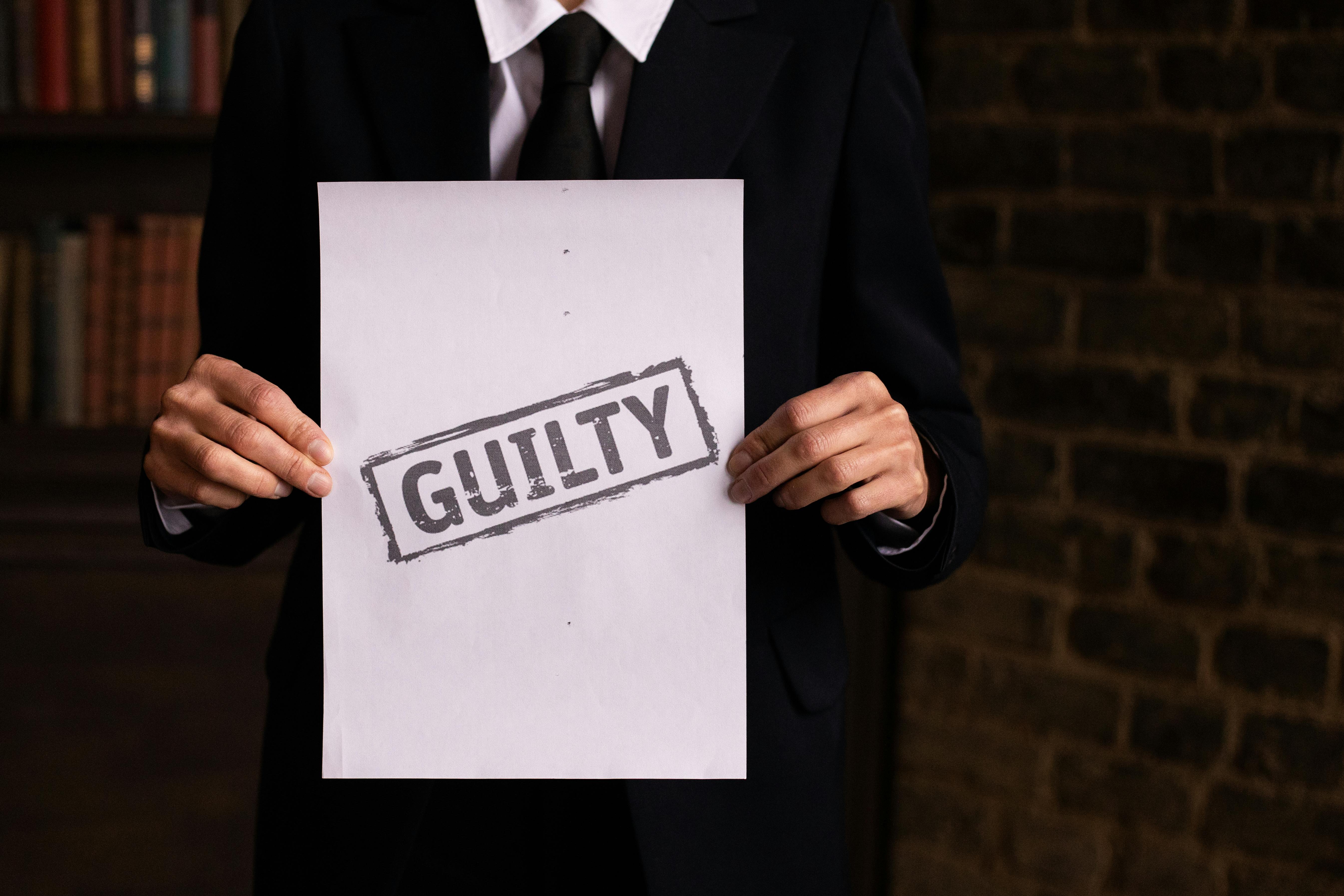 A businessman in a formal suit holding a sign with the word 'GUILTY.'