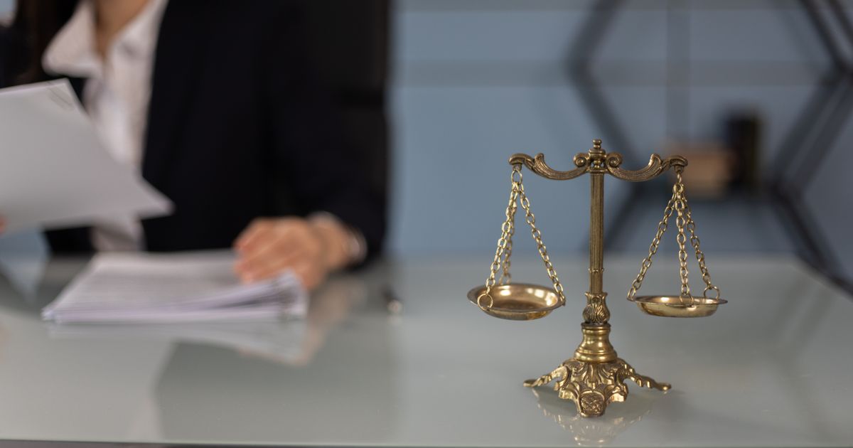 Brass balance scales on a glass desk, with a blurred person in a dark blazer paging through documents behind them.