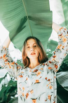 Young woman posing under large green leaves, wearing a floral dress in an outdoor setting.