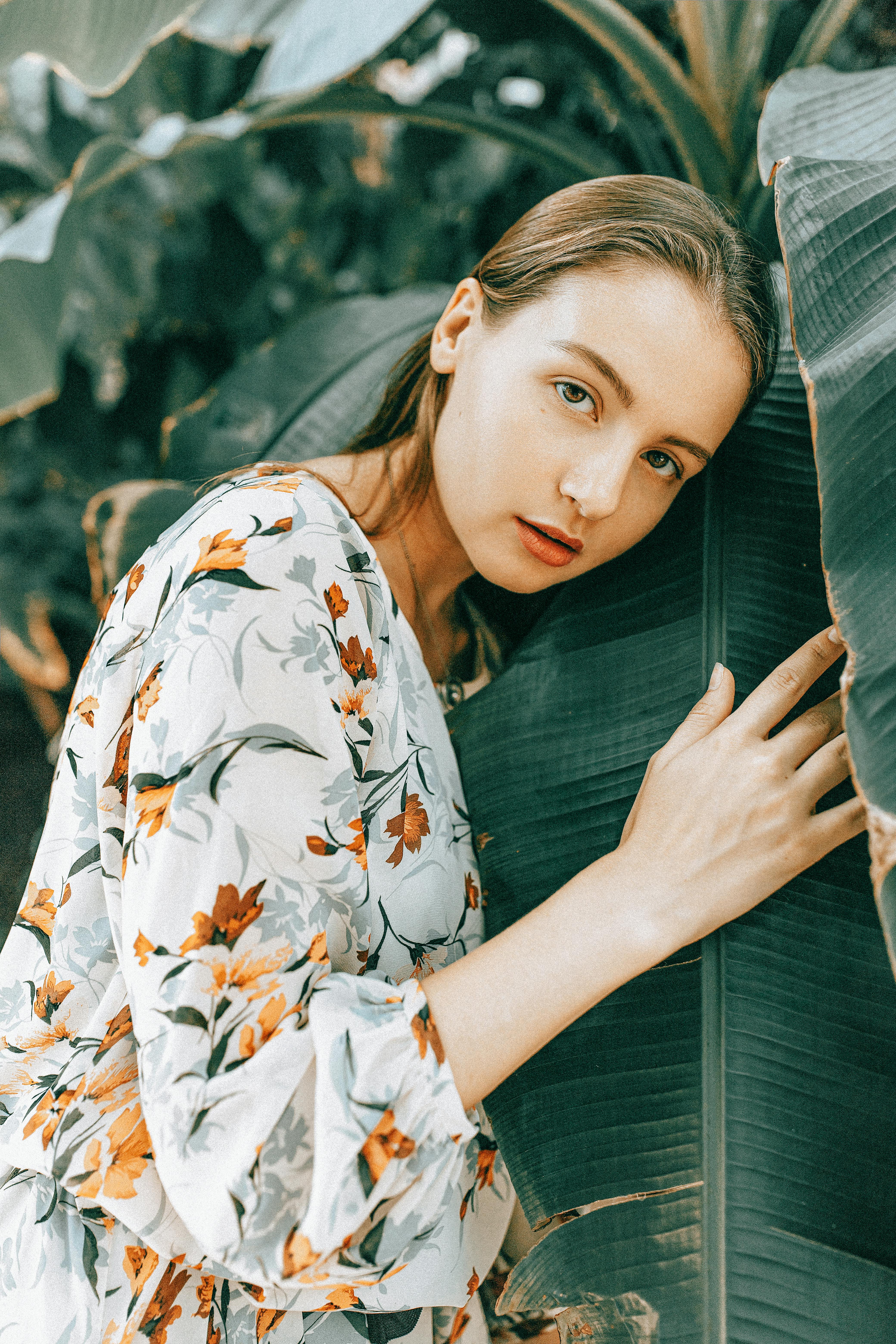 A woman in a floral dress embraces a large leaf in a serene outdoor setting.