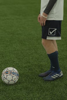 Soccer player standing on grass field with ball, wearing sports gear.