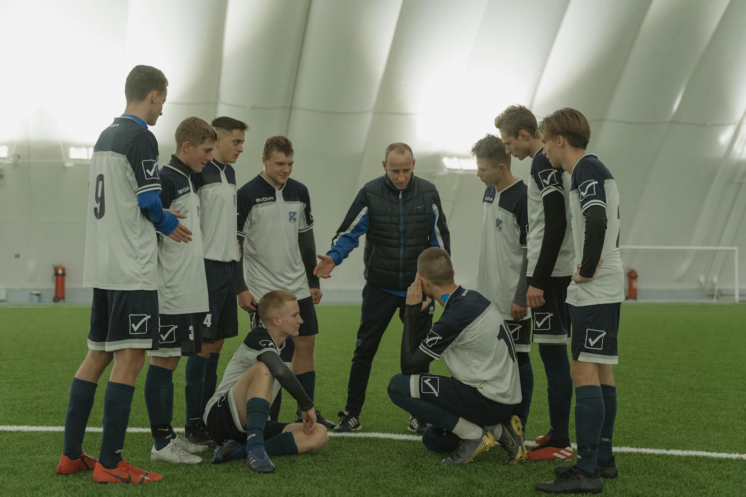 A soccer coach gives tactical advice to a youth team indoors on artificial turf.