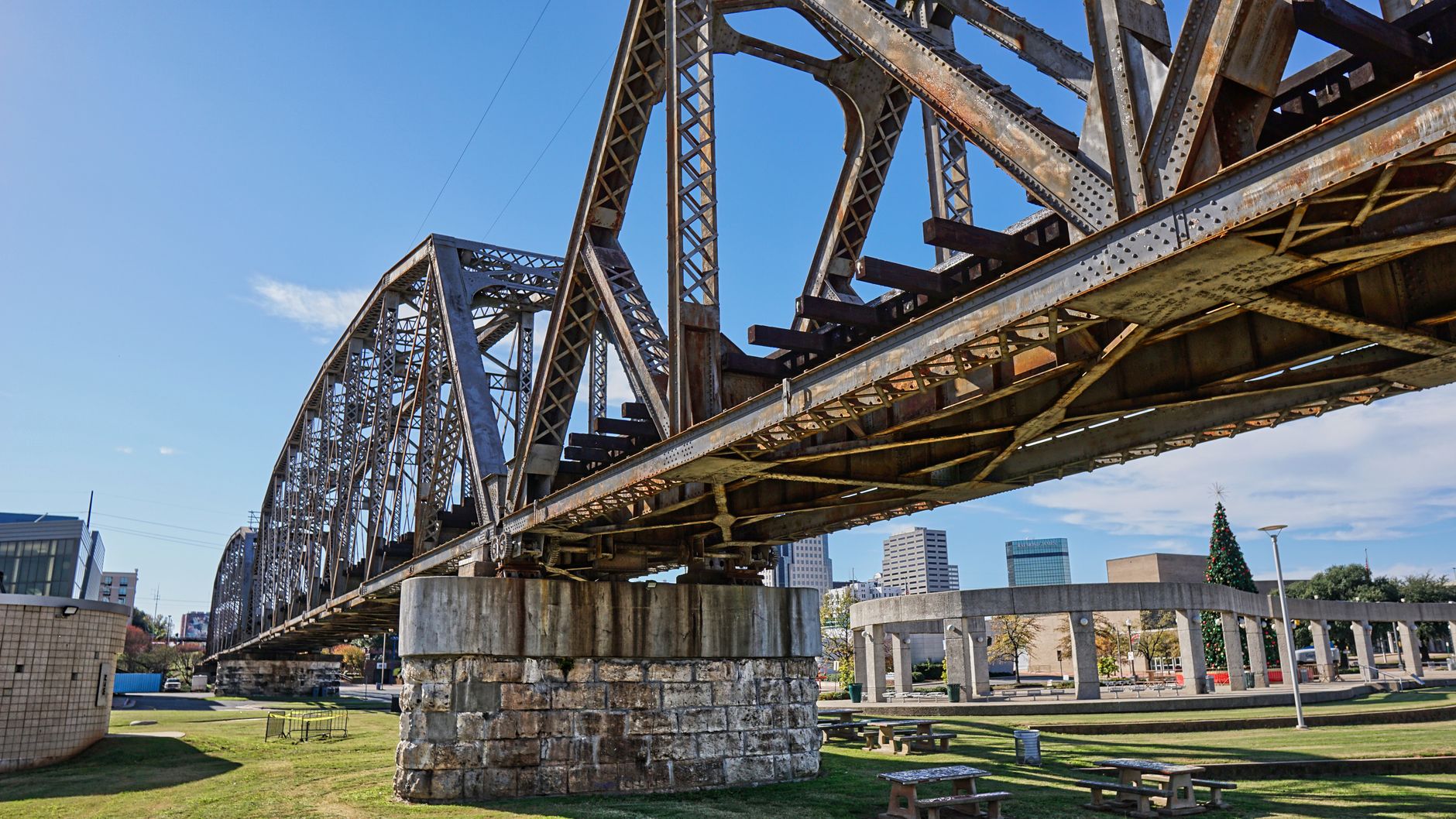 Rusty steel bridge in a Shreveport park against a clear blue sky, showcasing city skyline.
