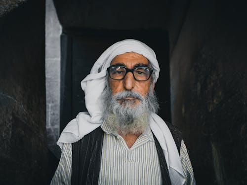 Senior ethnic male with gray beard in traditional apparel and headwear looking at camera between masonry construction walls