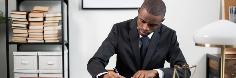 Businessman in formal suit writing notes at office desk with books and scales of justice.