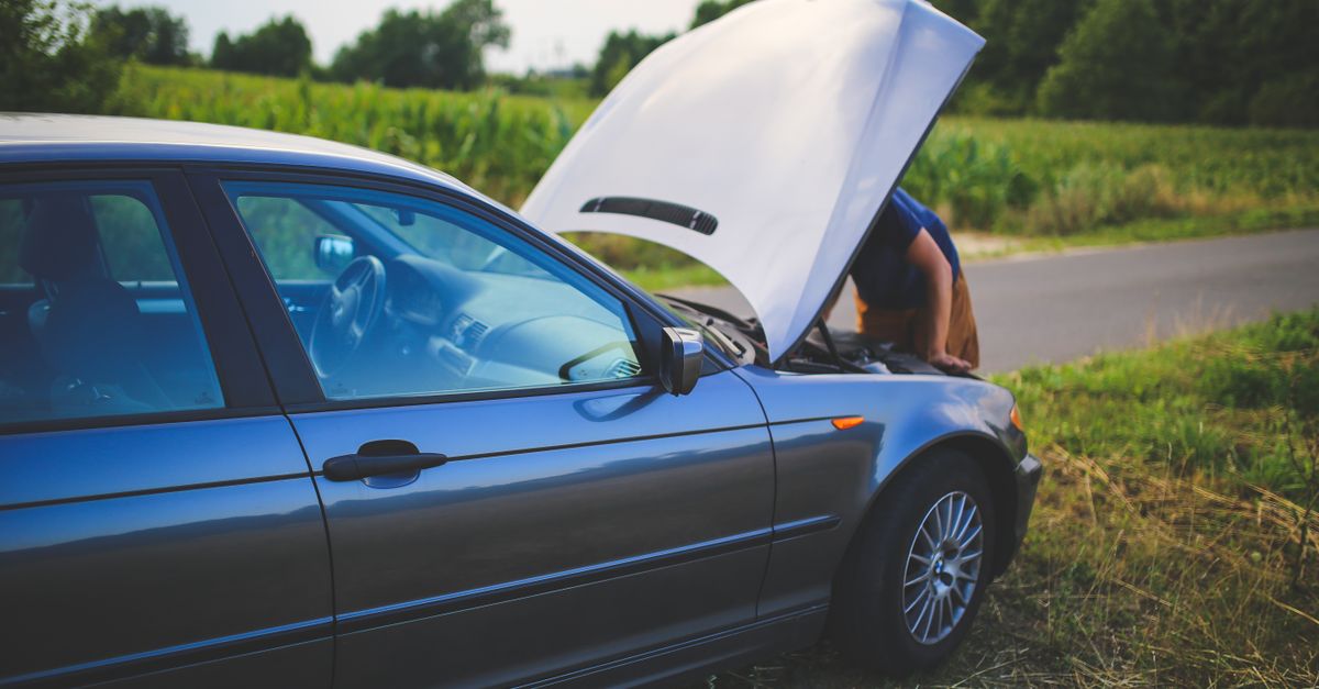 Man checking car engine with hood open by the side of a rural road.
