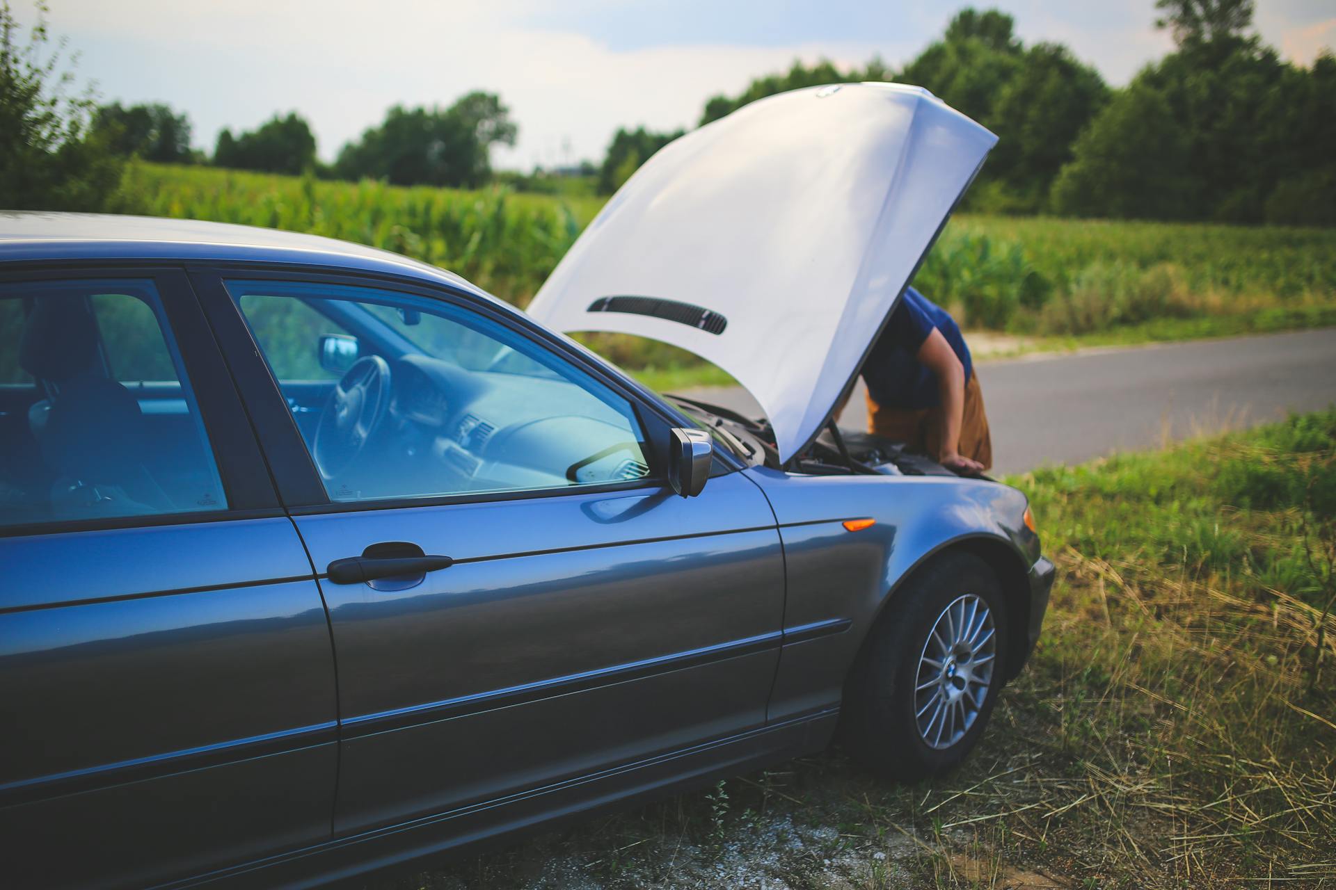 Man checking car engine with hood open by the side of a rural road.