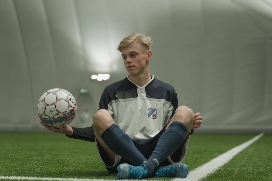Teen soccer player sitting on indoor field, holding ball in training facility.