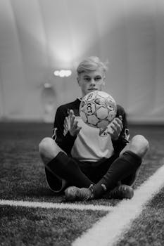 Black and white image of a young soccer player sitting with a soccer ball indoors on a field.