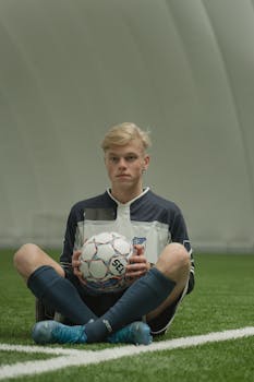 Teen soccer player in uniform sitting on artificial grass indoors, holding a soccer ball.