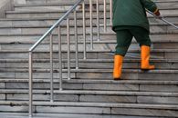 An individual in green attire and orange boots ascends marble stairs, holding a railing.