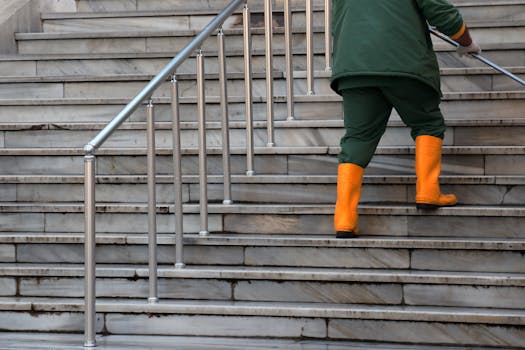 An individual in green attire and orange boots ascends marble stairs, holding a railing.