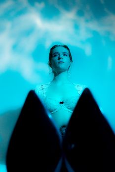 Through shoes tips young slim female in white bra sitting on floor against wall in dark room in blue light and looking away
